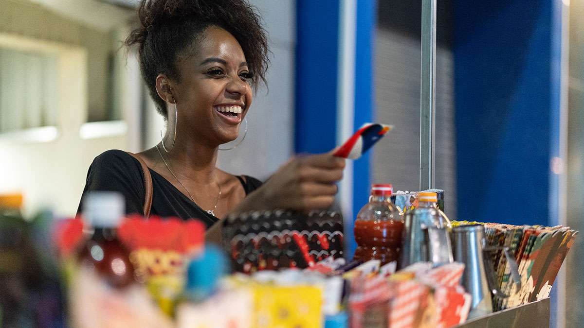 A young Brazilian woman handing over her British Airways Prepaid Mastercard to buy some items at a market while smiling and laughing with the vendor, out of shot.