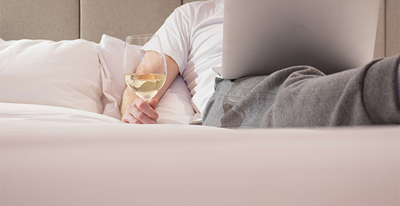 Young man lying on white bed, watching computer tablet and drinking a glass of white wine.
