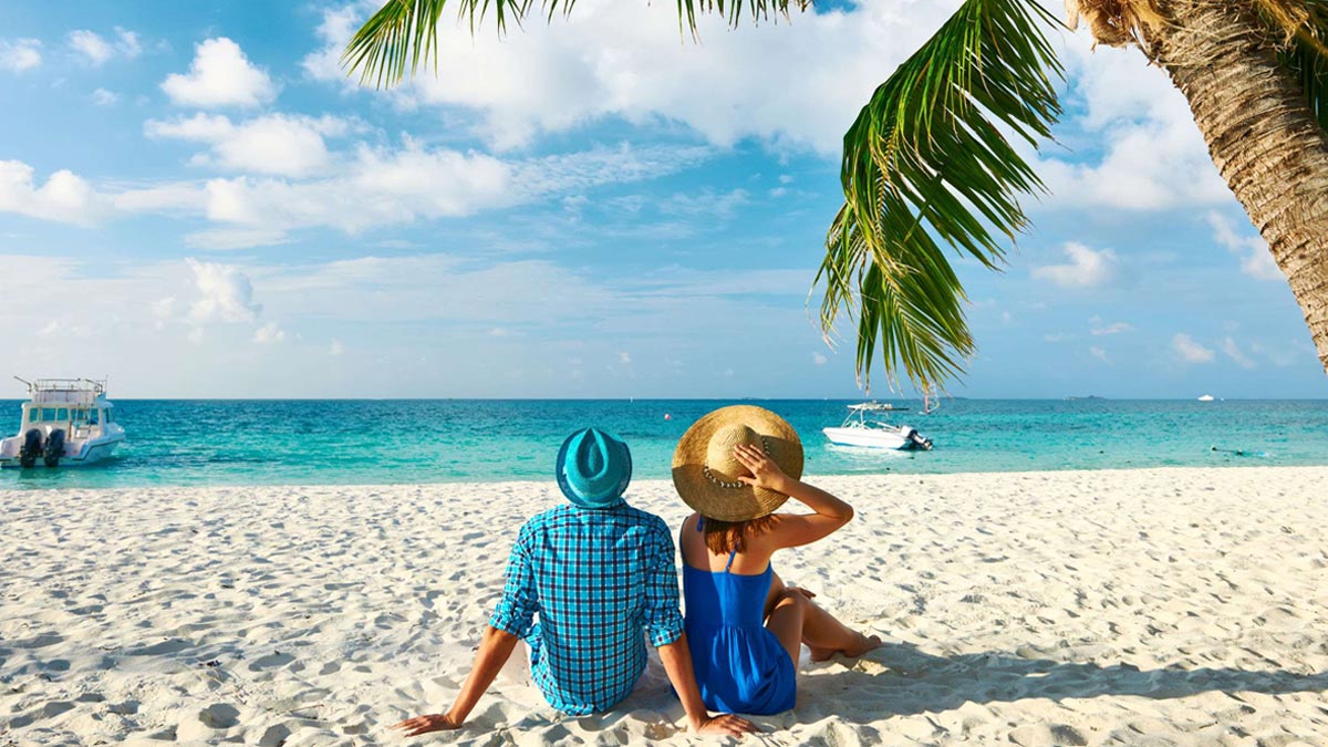 A couple wearing hats sat on a beach in the sun.