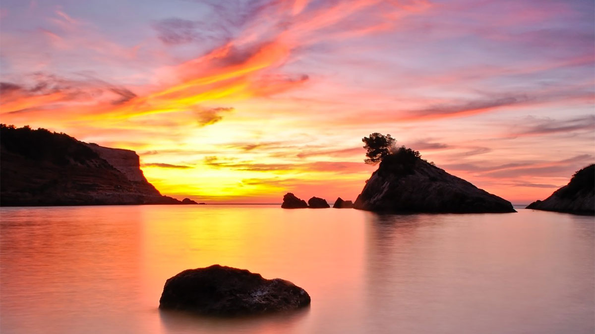 Sunset over sea with rock in water and mountain in background, Ibiza.