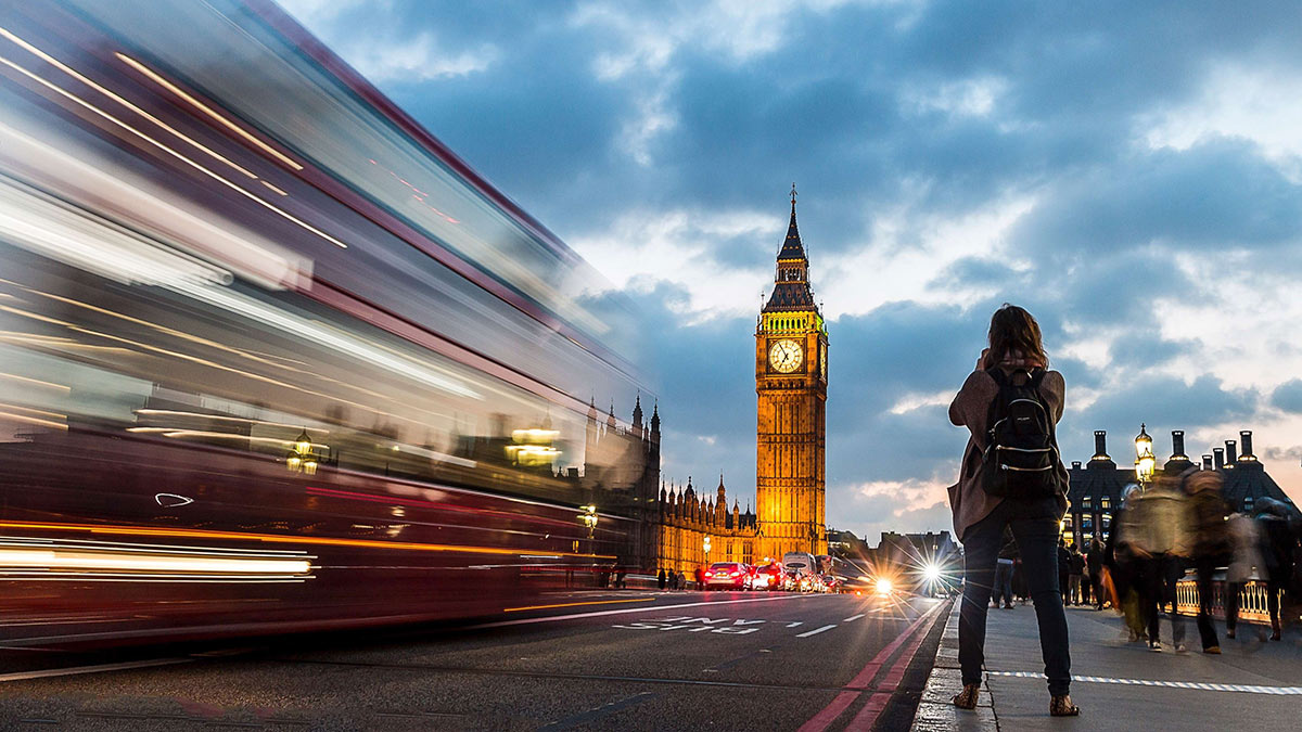 View of a woman standing on the foot path by a bus and Big Ben against a cloudy sky at sunset.