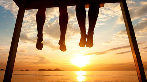 坐在日落海滩码头上的一对情侣的脚的剪影。