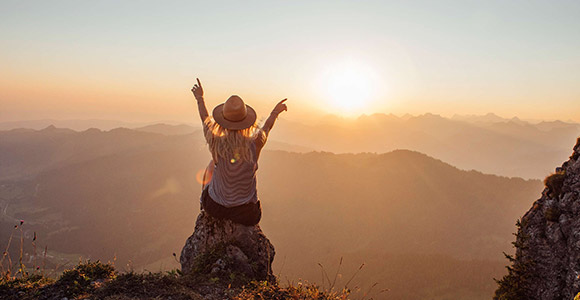Woman sitting on a rock on top of a hill looking into the horizon as the sun sets with arms in the air.